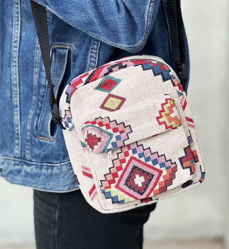 Puede incluir: Un peque&ntilde;o bolso bandolera beige con un estampado geom&eacute;trico colorido. El bolso tiene una correa ajustable negra y un cierre de cremallera.
