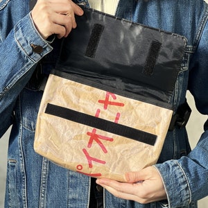 Może przedstawiać: Brązowa torba listonoszka z czarnymi paskami i klapką. Torba jest wykonana z materiału przypominającego papier pochodzący z recyklingu i ma nadrukowane czerwone japońskie znaki.