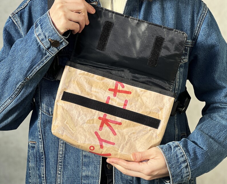 Może przedstawiać: Brązowa torba listonoszka z czarnymi paskami i klapką. Torba jest wykonana z materiału przypominającego papier pochodzący z recyklingu i ma nadrukowane czerwone japońskie znaki.