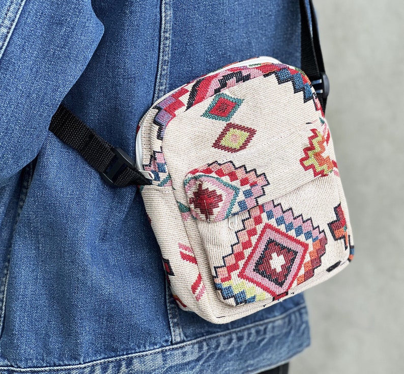 Puede incluir: Un peque&ntilde;o bolso bandolera beige con un estampado geom&eacute;trico colorido. El bolso tiene una cremallera y una correa ajustable.
