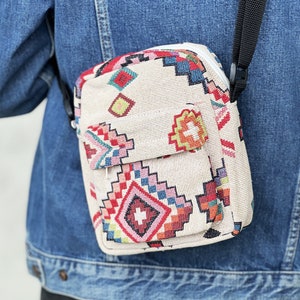 Puede incluir: Un peque&ntilde;o bolso bandolera beige con un estampado geom&eacute;trico colorido. El bolso tiene cierre de cremallera y una correa negra ajustable.