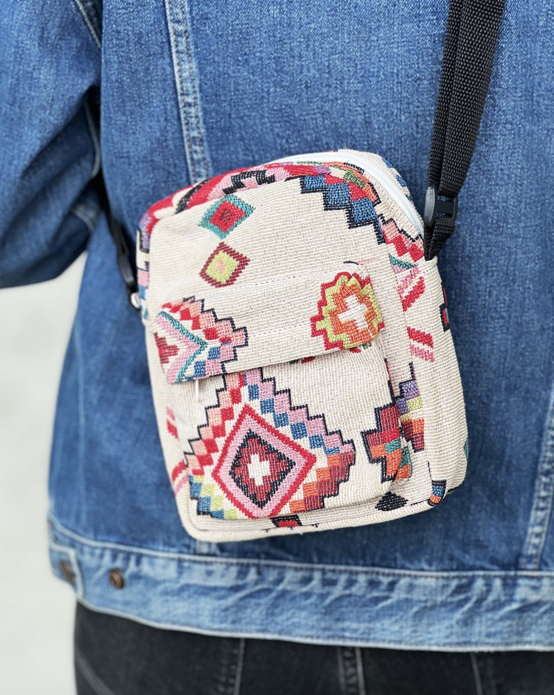 Puede incluir: Un peque&ntilde;o bolso bandolera beige con un estampado geom&eacute;trico colorido. El bolso tiene cierre de cremallera y una correa negra ajustable.