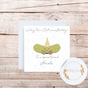 Peut inclure: Carte de vœux avec un cornichon de dessin animé portant un chapeau de fête. La carte dit "Wishing You a Dill-icious Birthday!" et "To a special friend Amelie." Un autocollant rond avec des feuilles d'or et du texte est en bas à droite.