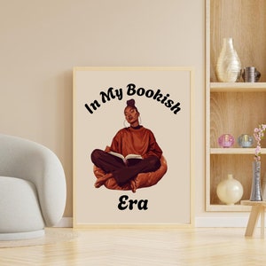 Op de afbeelding: Een ingelijst afdruk met een vrouw die op een stoel zit en een boek leest. De tekst op de afdruk luidt "In My Bookish Era".