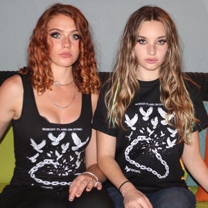 May include: Two people wearing black tops with the text "NOBODY PLANS ON DYING" and a graphic of birds and a chain. One top is a tank top, the other is a t-shirt. The background includes a green and orange cushion.