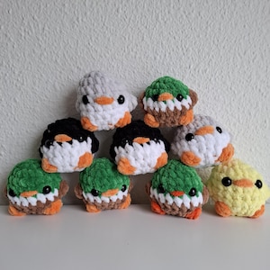 以下が含まれることがあります： さまざまな色の手作りのかぎ針編みのあひるのコレクション。アヒルは、オレンジ色のくちばしと足を持つ、白、黒、グレー、緑、黄色の体を持っています。これらのぬいぐるみは白い表面に配置されています。