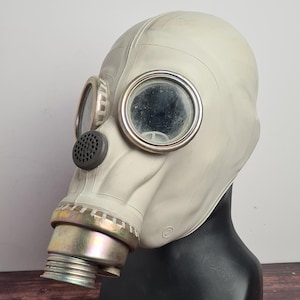 Puede incluir: Una máscara de gas vintage gris claro, con grandes oculares circulares y un filtro circular. La máscara se muestra sobre una cabeza de maniquí negra, sobre un fondo neutro.