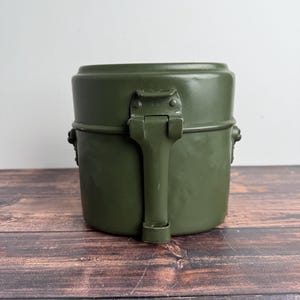 以下が含まれることがあります： オリーブグリーンの金属製メスキット。ヒンジ付きのハンドルと安全な留め金が付いています。キットは円筒形で、上部と下部が丸みを帯びています。ハンドルは折りたたまれ、木製の表面に置かれています。