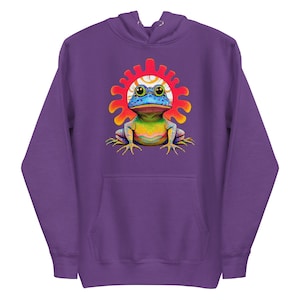 May include: A purple hoodie featuring a colorful frog illustration. The frog has a blue head, yellow eyes, and a rainbow body, set against a red and white sunburst design. The hoodie has a front pocket and a drawstring hood.
