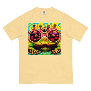 May include: A light yellow t-shirt featuring a colorful, psychedelic frog illustration. The frog has large, multi-colored eyes and a green body. The background of the graphic is blue and yellow.