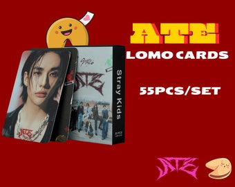 Puede incluir: Un conjunto de 55 tarjetas de fotos de Stray Kids con una foto en blanco y negro de un miembro del grupo. Las tarjetas están empaquetadas en una caja con el nombre del grupo y el texto "Lomo Cards" impreso en ella. La caja está decorada con un gráfico de galleta de la fortuna.