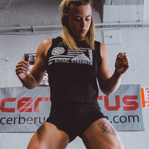 May include: A woman wearing black shorts and a black tank top with the text "Ritual Strength" is lifting a barbell. She is wearing black socks and black shoes. The barbell is resting on weight plates. The background is a white brick wall with a sign that says "Cerberus.com".