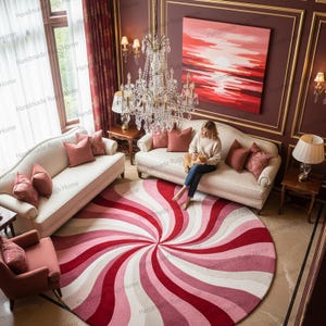 May include: A round rug with a spiral design in shades of red, pink, and white dominates the room. The rug is placed in front of a cream-colored sofa. The room also features a chandelier and a painting of a sunset.