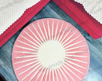 May include: A pink and white round rug with a sunburst design. The rug is made of soft, fluffy material and has a white center circle.