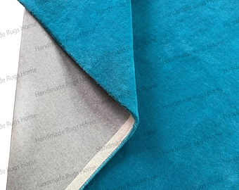Puede incluir: Primer plano de una alfombra turquesa con reverso gris. La tela turquesa está doblada, revelando el reverso gris y un borde blanco. La alfombra tiene una textura suave y afelpada.
