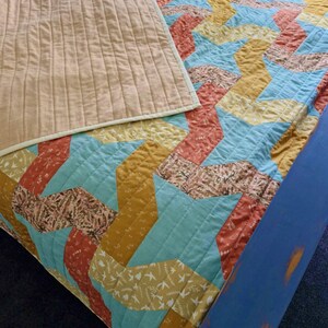 May include: A colourful quilt with a geometric pattern in shades of blue, orange, yellow, and brown. The quilt has a tan backing with vertical stitching. The quilt is folded over a blue bolster.