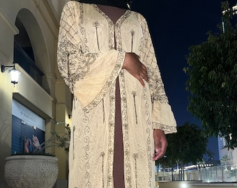 Op de afbeelding: Een lange, doorschijnende, beige cardigan met gouden borduurwerk en kralenfranjes. De cardigan heeft een V-hals en lange mouwen.