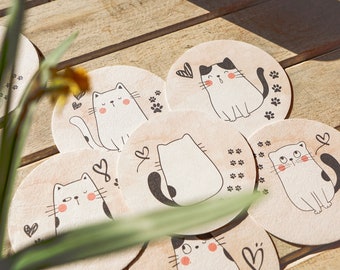 May include: Set of eight round wooden coasters featuring a cute cat design. Each coaster has a different cat illustration with black and white details and pink cheeks. The coasters are perfect for protecting surfaces from heat and spills.