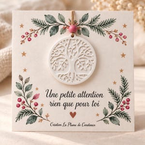 Peut inclure: Un ornement circulaire blanc représentant l'arbre de vie est suspendu par une ficelle avec une perle rose sur une carte carrée. La carte présente des illustrations aquarelles de feuilles vertes, baies rouges et étoiles dorées, avec le texte "Une petite attention rien que pour toi" et "Création La Plume de Constance" imprimés en noir.