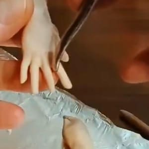 Può includere: Un tutorial sulla scultura di mani realistiche con argilla polimerica. L'immagine mostra un primo piano di un paio di mani fatte di argilla polimerica, con uno strumento che manipola l'argilla. Il testo "Polymer Clay Tutorial Sculpting Realistic Hands Video Lesson" è visibile.