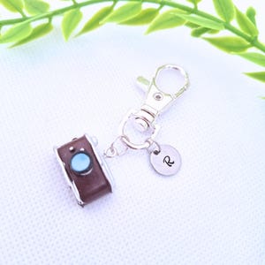 May include: A brown camera charm with a blue lens and silver accents is attached to a silver keychain. A small, round charm with the letter "R" is also attached. The background is white with green leaves.