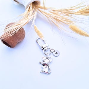 Op de afbeelding: Een zilverkleurige muissleutelhanger met een karabijnhaak en een kleine ronde bedel met de letter "V". De sleutelhanger wordt gepresenteerd op een wit oppervlak met gedroogde tarwestengels en een bruin decoratief object.