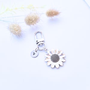 May include: A silver-coloured keyring with a daisy flower charm, featuring a dark centre and a small, round tag with the number 2. The keyring includes a clasp for easy attachment.