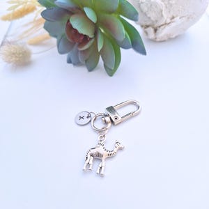 Op de afbeelding: Een zilverkleurige sleutelhanger met een kameelbedel, een ronde tag met de letter "X" en een sluiting. De sleutelhanger ligt op een wit oppervlak met een vetplant en een steen op de achtergrond.