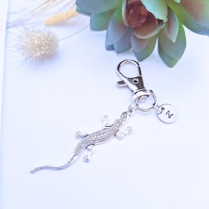 May include: Silver lizard keyring with a lobster clasp and a small round charm with the letter "N". The lizard charm has detailed scales. The keyring is on a white surface with a succulent and dried plants in the background.