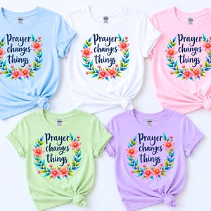 Peut inclure: Cinq t-shirts à manches courtes et col rond en bleu clair, blanc, rose clair, vert clair et lavande. Chaque t-shirt présente un motif central de couronne florale rose, orange et verte entourant le texte bleu foncé Prayer changes things.