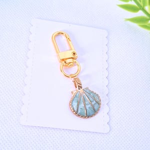 May include: A gold-coloured keyring with a seashell charm. The charm is a light blue colour with gold-coloured trim. The keyring is attached to a white card with a scalloped edge.