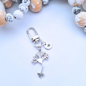 May include: Silver neuron charm keyring with a lobster clasp and a small round tag with the letter "w". The neuron charm has a detailed, tree-like design. The keyring is on a white background.