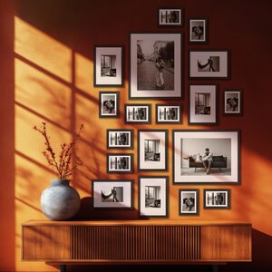 Peut inclure: Une composition murale de cadres noirs contenant des photographies en noir et blanc. Les cadres sont accrochés sur un mur orange au-dessus d'un meuble en bois. Un vase avec des branches est posé sur le meuble.