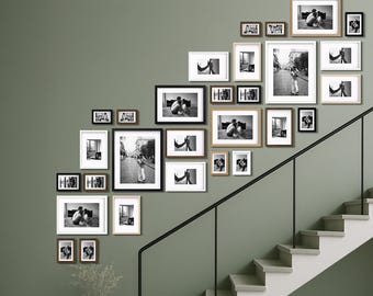 May include: A gallery wall arrangement of framed black and white photographs in various sizes and frame styles. The frames are arranged on a green wall near a staircase. The photographs depict people and scenes.