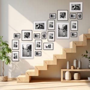 Könnte beinhalten: Eine Galeriewand mit gerahmten Schwarz-Weiß-Fotografien in verschiedenen Größen. Die Rahmen sind schwarz mit weißen Passepartouts, was eine zusammenhängende Darstellung ergibt. Die Anordnung befindet sich an einer hellen Wand, mit einer Holztreppe und dekorativen Elementen im Hintergrund.
