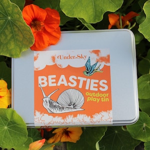 May include: A silver metal tin with a colorful label that says "BEASTIES outdoor play tin" and features a snail and a bluebird. The tin is surrounded by green leaves and orange flowers.