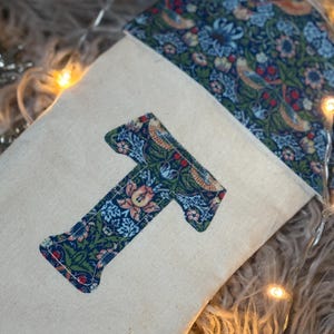 May include: A cream-coloured Christmas stocking with a floral patterned cuff and a large letter "T" appliqué. The "T" is made of the same floral fabric, featuring blue, green, and peach tones. The background is a soft, neutral colour.