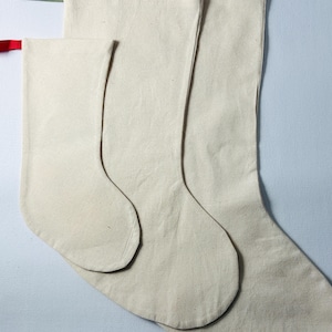 May include: Three plain, off-white canvas stockings of varying sizes arranged in a vertical stack. Each stocking features a simple, unadorned design with a clean, neutral fabric texture and a small fabric hanging loop attached to the top corner.