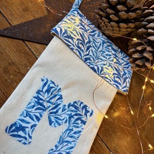 May include: Cream-colored Christmas stocking with a blue and white floral pattern on the cuff and the letter "M". The stocking is decorated with string lights and pine cones, creating a festive holiday theme.