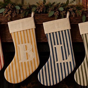 May include: Four Christmas stockings hanging in a row, each featuring vertical stripes in red, yellow, blue, and green. Every stocking has a cream-colored cuff at the top and a large, centered, cream-colored capital letter: C, B, L, and A.