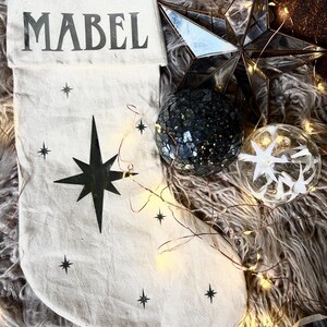 May include: A cream-colored Christmas stocking with the name "MABEL" printed in black. The stocking features a large black star and smaller star accents. Decorative ornaments and a star-shaped decoration are also visible.