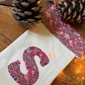 Könnte beinhalten: Ein cremefarbener Weihnachtsstrumpf mit einem Applikation des Buchstaben "S" mit rotem Blumenmuster. Eine passende Schlaufe mit rotem Blumenmuster ist angebracht. Zwei Tannenzapfen und ein sternförmiger Schmuck im Hintergrund.