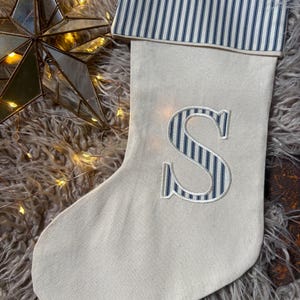 May include: A cream-coloured fabric Christmas stocking featuring a cuff with a navy blue and white vertical pinstripe pattern. The centre of the stocking displays a large, appliqued letter S in the same navy blue and white pinstripe fabric.