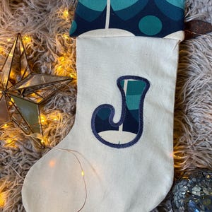 May include: A cream-coloured fabric Christmas stocking featuring a cuff with a teal, navy, and white geometric pattern. The centre of the stocking displays a large, appliqued letter J in the same teal, navy, and white geometric fabric design.