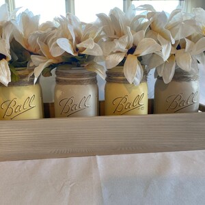 Könnte beinhalten: Vier rustikale Einmachgläser mit einem Juteband und weißen Blumen sind in einer Holzkiste angeordnet. Die Gläser sind hellgelb lackiert und haben das Wort "Ball" auf der Vorderseite geprägt.