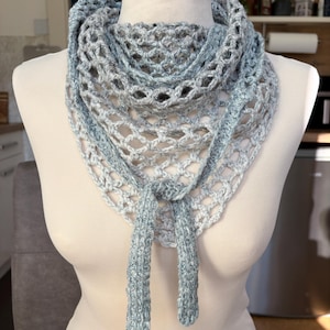 May include: A handmade, open-weave crochet scarf in shades of blue and gray. The scarf is draped on a mannequin, showcasing its triangular shape and textured design. The scarf has two long, knotted ties at the front.
