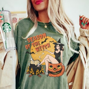 May include: Green t-shirt featuring a vintage-style graphic of a witch sitting on a pumpkin. The graphic reads "Season of the Witch".
