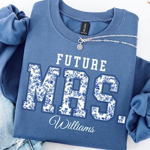 May include: Indigo blue sweatshirt with the text "FUTURE MRS." in white block letters with a blue floral pattern. The name "Williams" is scripted below. A silver necklace with a pendant rests on the sweatshirt. The sweatshirt is folded on top of a pair of blue jeans.