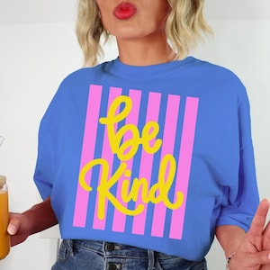May include: A blue t-shirt with the words "be Kind" in yellow script, set against a pink striped background. The shirt is paired with denim shorts. A glass with a bamboo lid and a straw is held in the hand.