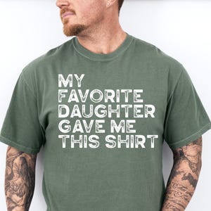 Pu&ograve; includere: Una maglietta verde muschio con la scritta "MY FAVORITE DAUGHTER GAVE ME THIS SHIRT" in lettere bianche. La maglietta &egrave; di colore pieno e ha maniche corte. La maglietta &egrave; indossata da una persona.